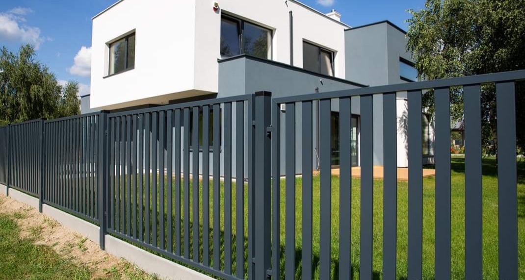 Nowoczesne ogrodzenie z metalowych sztachet w kolorze grafitowym, z widoczną betonową podmurówką i minimalistycznym domem w tle.