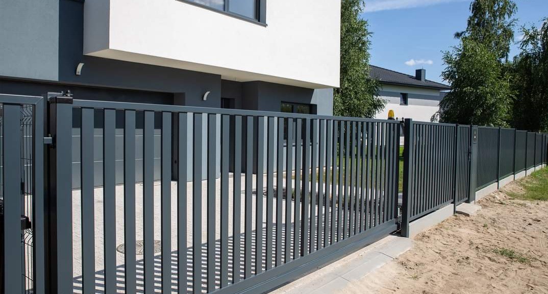 Nowoczesna, przesuwna brama i ogrodzenie z pionowych, metalowych profili w odcieniach szarości, dopasowane do kolorystyki domu w tle.