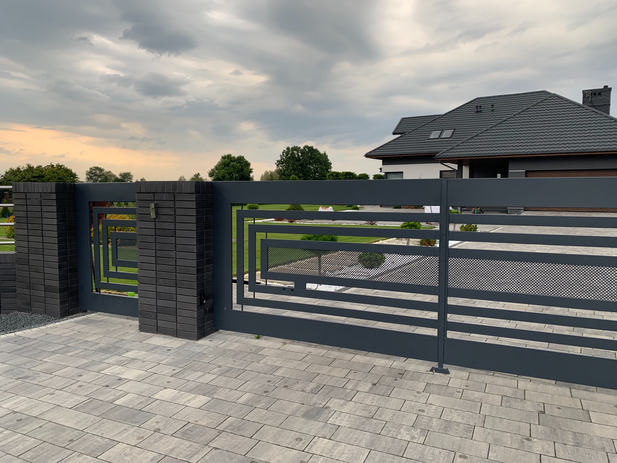Nowoczesna, przesuwna brama metalowa w kolorze grafitowym z geometrycznym wzorem, osadzona na murowanych słupkach z cegły klinkierowej, na tle domu z ciemną dachówką i pochmurnego nieba.