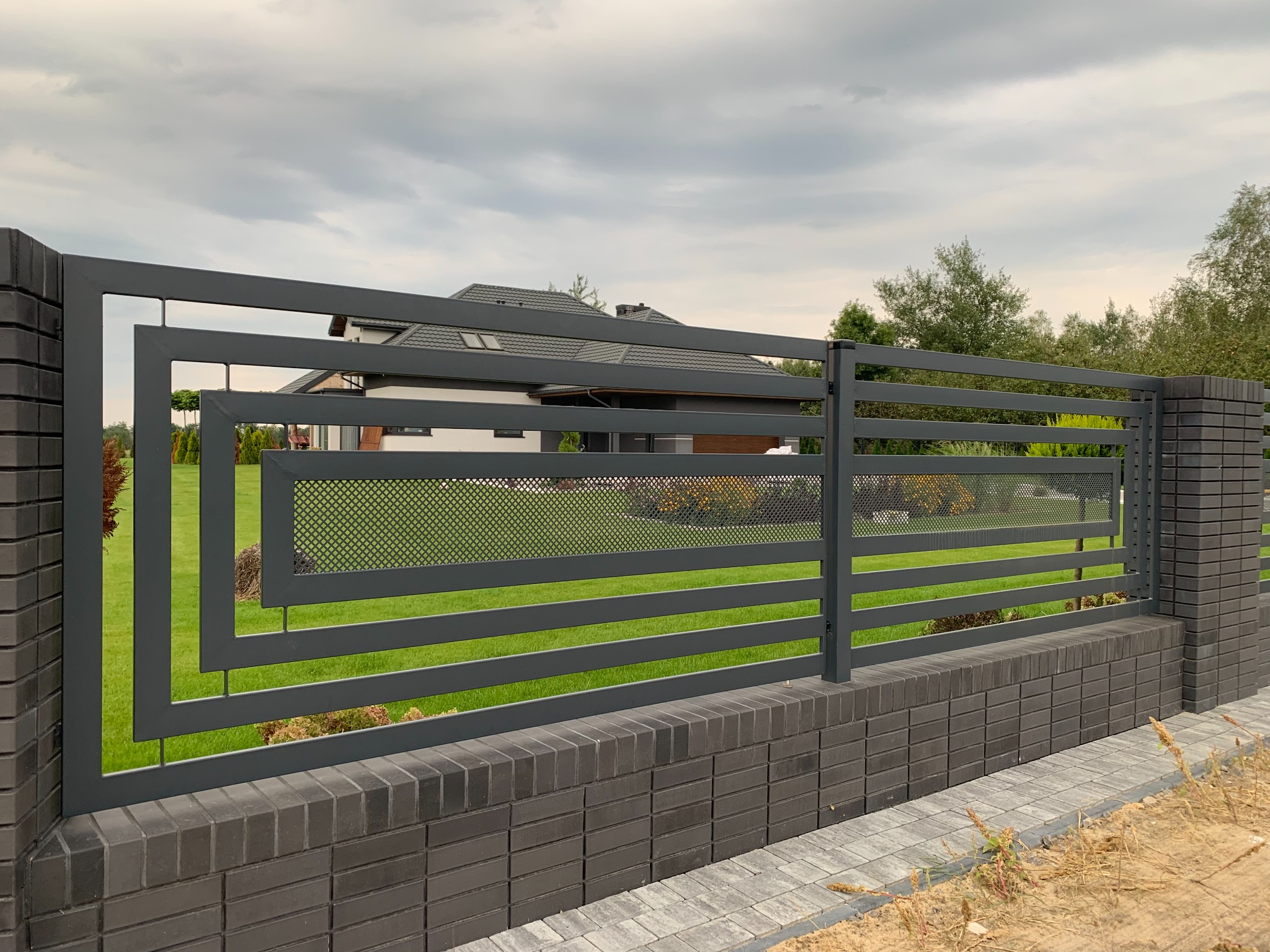 Nowoczesne, poziome przęsła w grafitowym kolorze z elementami ażurowymi, osadzone na murku z ciemnej cegły klinkierowej, z domem w tle.