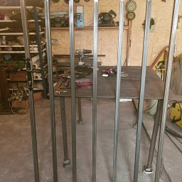 Kanak Welder - Surowa, metalowa rama bramy wjazdowej z zamontowanym zamkiem i klamką, ustawiona w warsztacie na tle stołu spawalniczego z narzędziami.