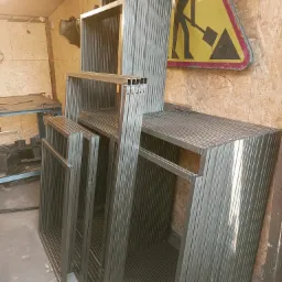 Stos metalowych ram z profili kwadratowych, przygotowanych do dalszej obróbki, w tle płyta OSB i znak drogowy ostrzegający o robotach.