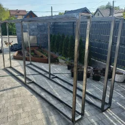 Metalowa konstrukcja szkieletowa, prawdopodobnie pod szklarnię, leżąca na kostce brukowej w ogrodzie, z widocznymi grządkami i tujami w tle.