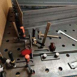 Kanak Welder - Przygotowane do spawania elementy metalowe, zamocowane na perforowanym stole spawalniczym za pomocą ścisków i kątowników magnetycznych, widoczne narzędzia pomiarowe i młotek.