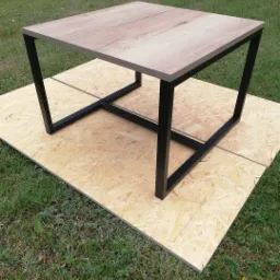 Kwadratowy stolik kawowy z blatem imitującym drewno i czarną metalową podstawą, ustawiony na płycie OSB na trawie.