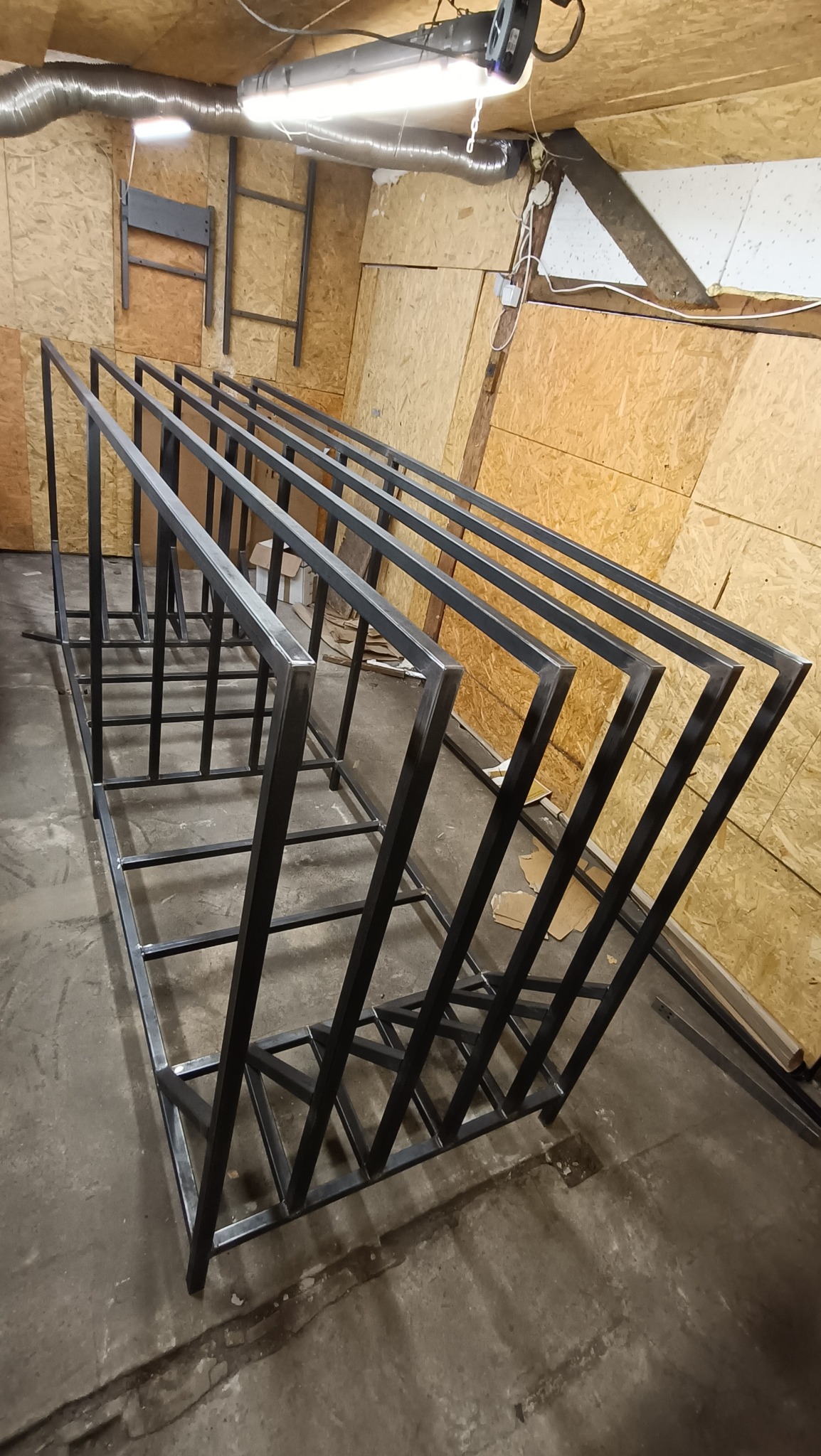 Surowe, metalowe ramy konstrukcji ustawione w warsztacie, widoczne ślady spawania i obróbki, surowa podłoga i ściany z płyt OSB w tle.