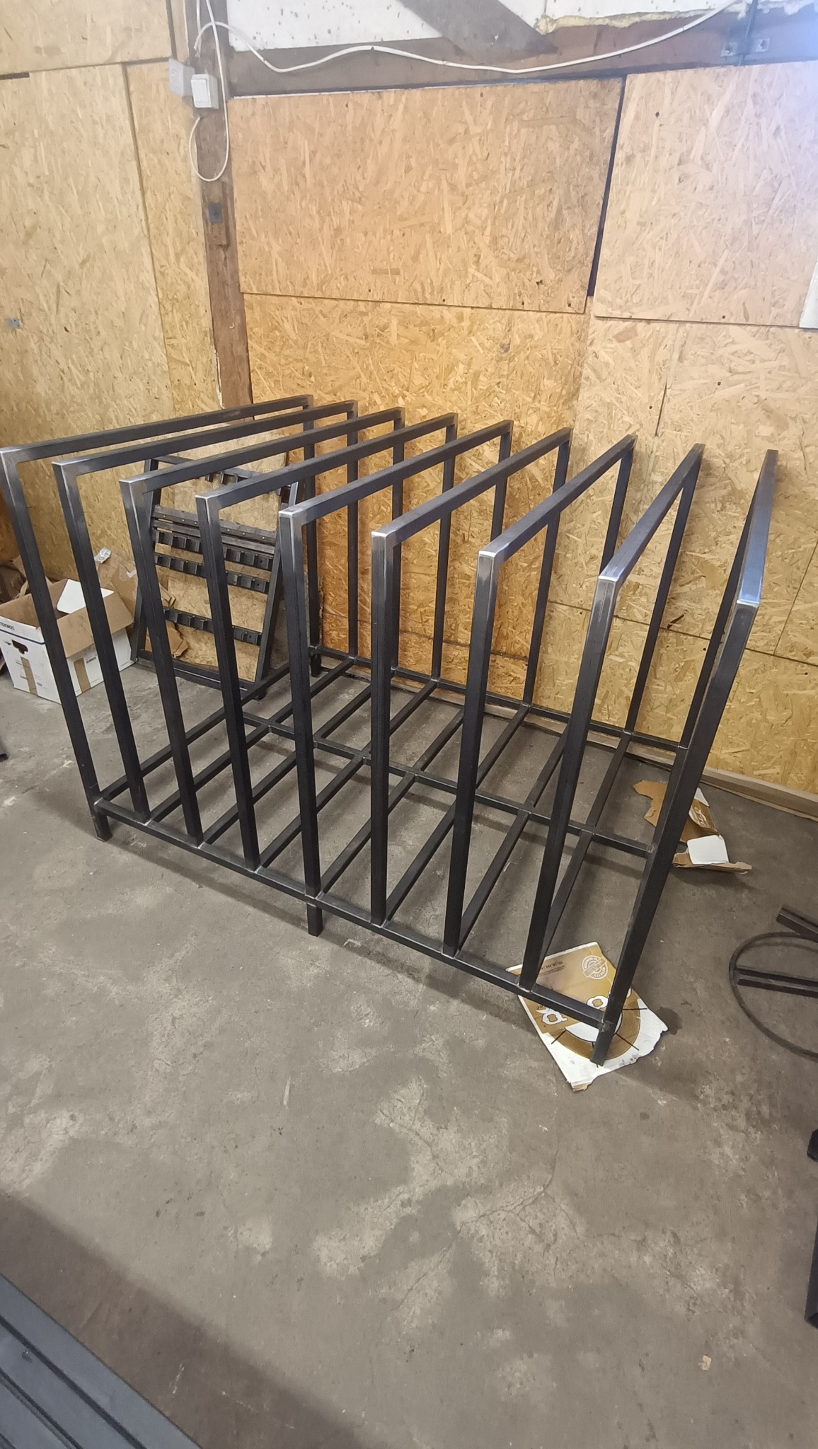 Surowa, metalowa konstrukcja z profili kwadratowych, prawdopodobnie regał lub stojak, w warsztacie na tle płyty OSB.