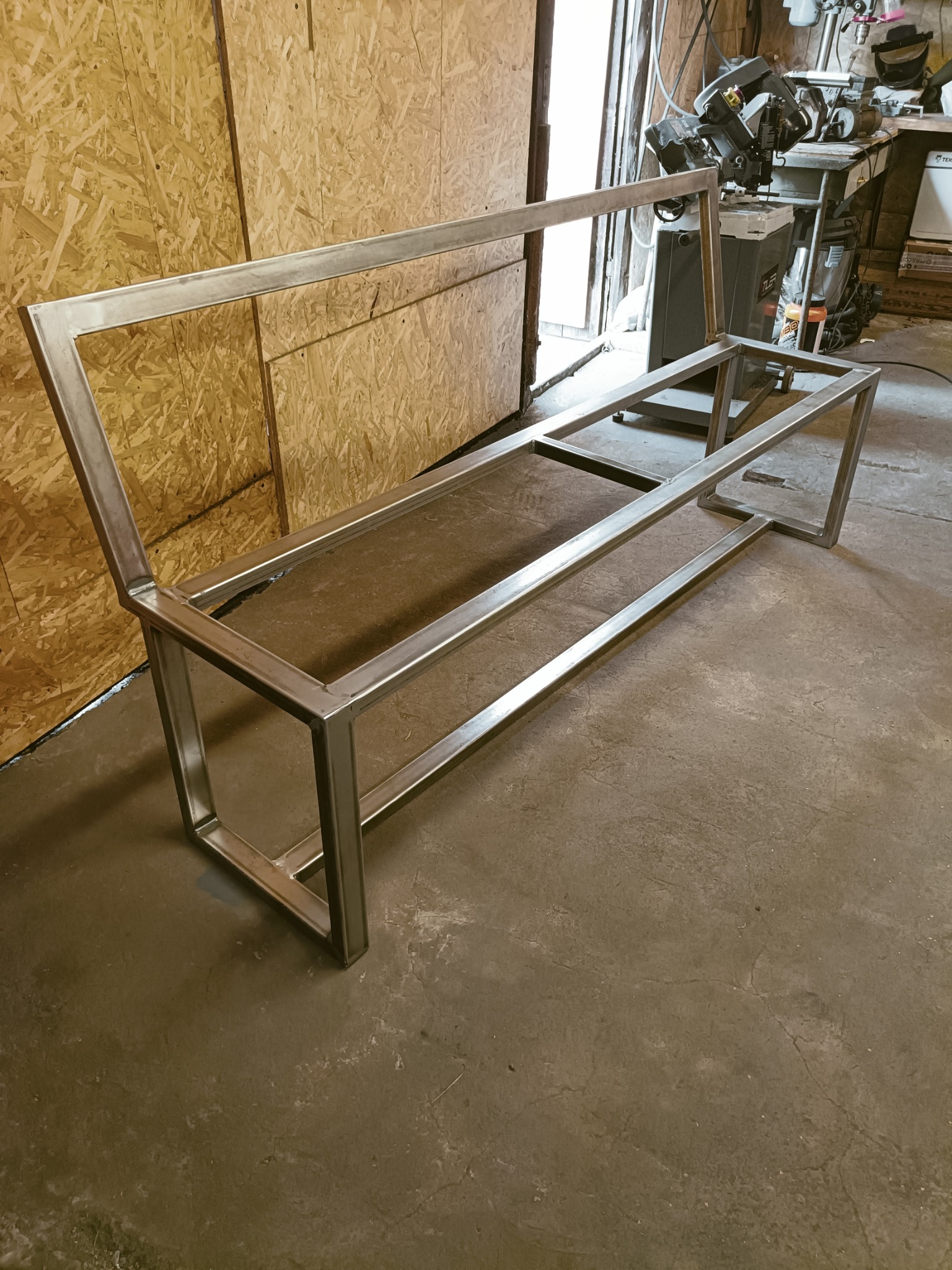 Surowy, spawany szkielet metalowej ławki, widoczny w warsztacie na tle ściany z płyt OSB i sprzętu spawalniczego.