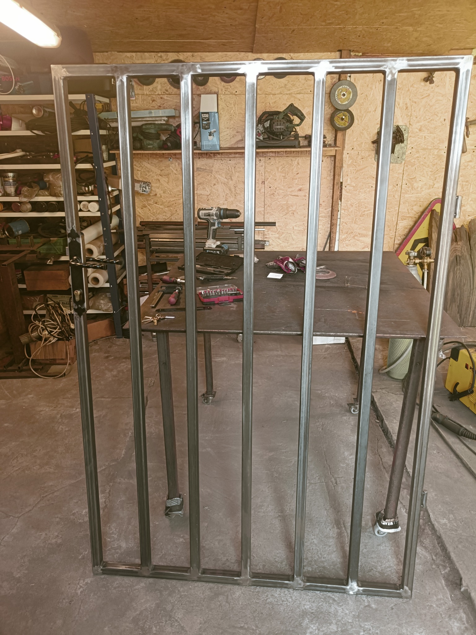 Surowa, metalowa rama bramy wjazdowej z zamontowanym zamkiem i klamką, ustawiona w warsztacie na tle stołu spawalniczego z narzędziami.