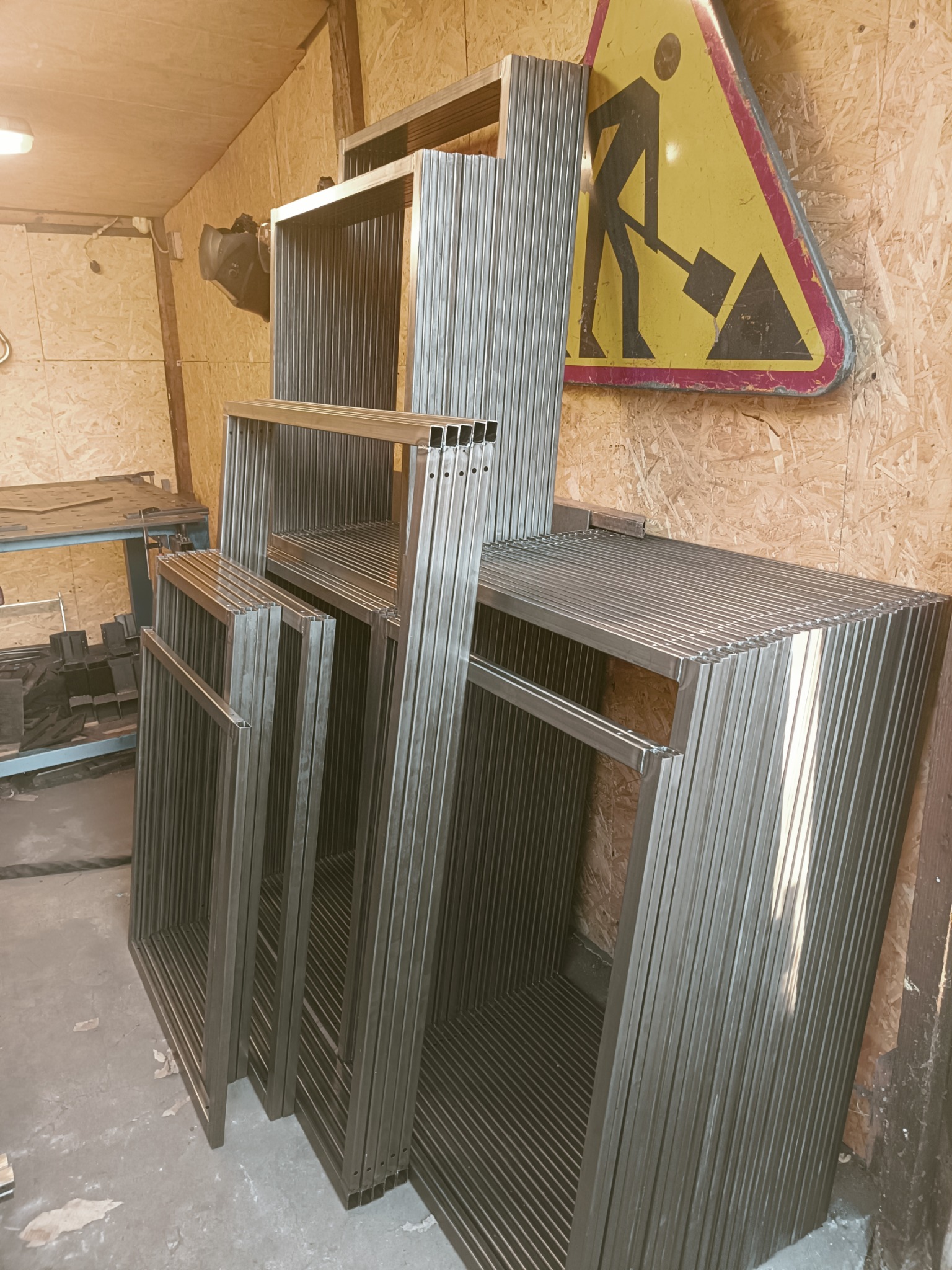 Stos metalowych ram z profili kwadratowych, przygotowanych do dalszej obróbki, w tle płyta OSB i znak drogowy ostrzegający o robotach.