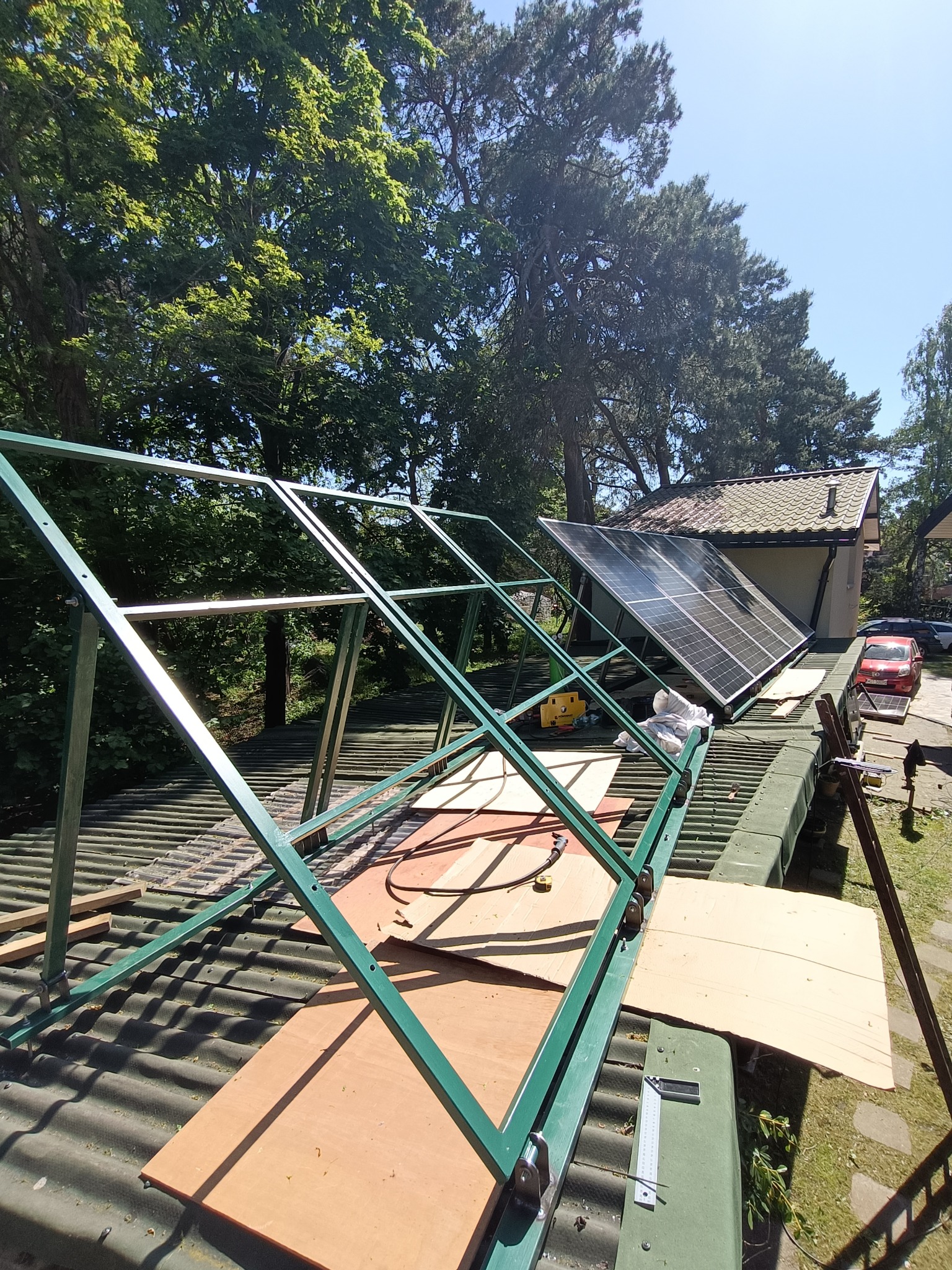 Montaż paneli słonecznych na dachu: zielona metalowa konstrukcja wspornikowa, panele fotowoltaiczne, narzędzia pomiarowe i fragment dachu domu w słoneczny dzień.