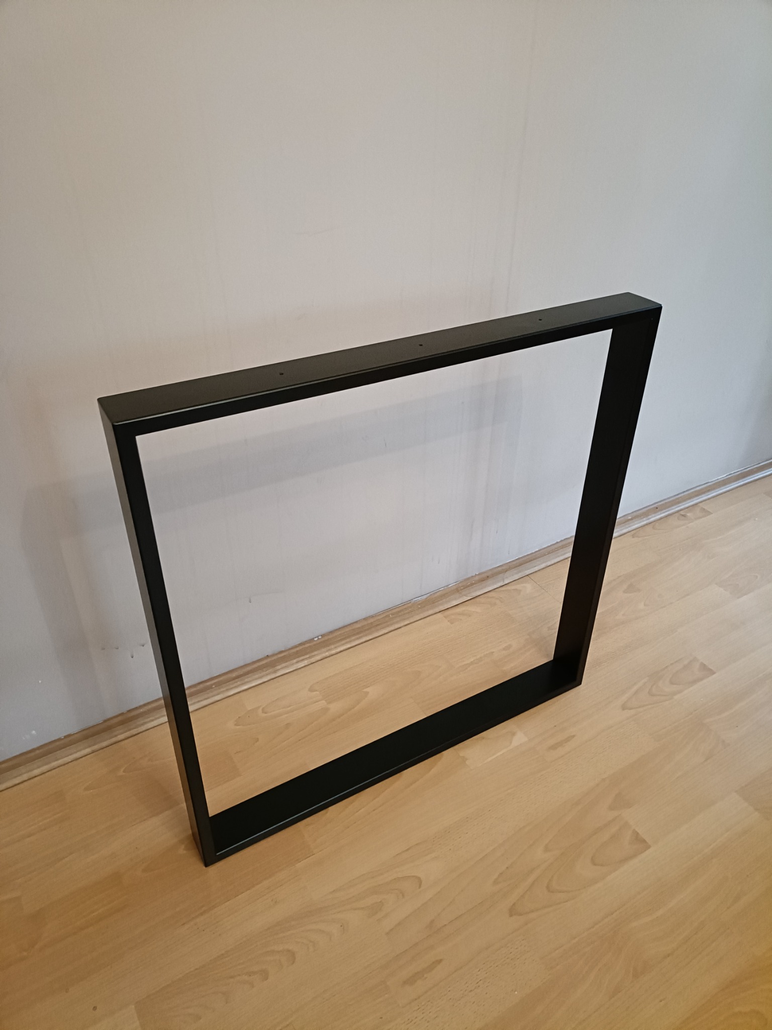 Metalowa rama w kształcie prostokąta, pomalowana na czarno, z otworami montażowymi, oparta o ścianę na drewnianej podłodze.
