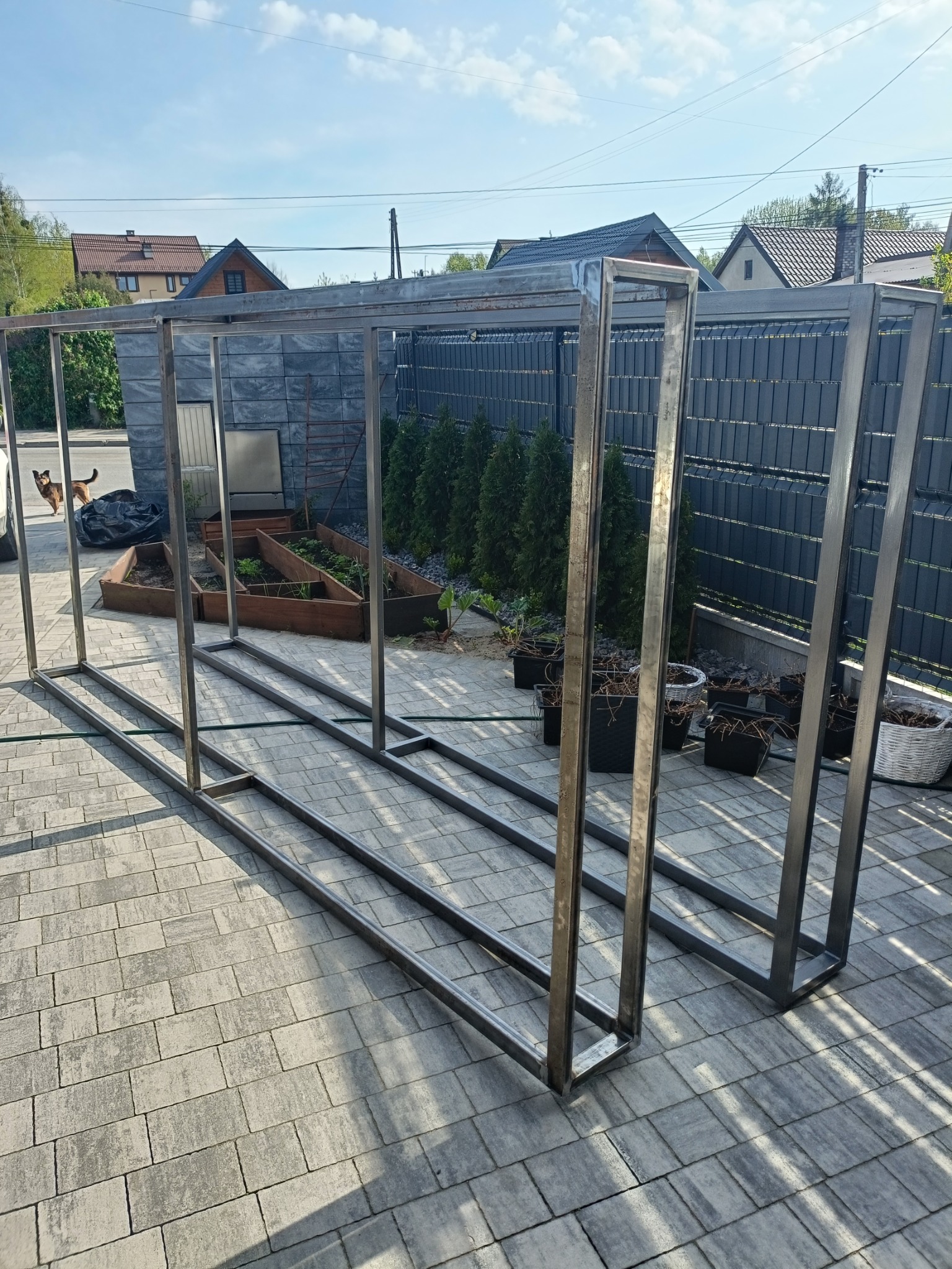 Metalowa konstrukcja szkieletowa, prawdopodobnie pod szklarnię, leżąca na kostce brukowej w ogrodzie, z widocznymi grządkami i tujami w tle.