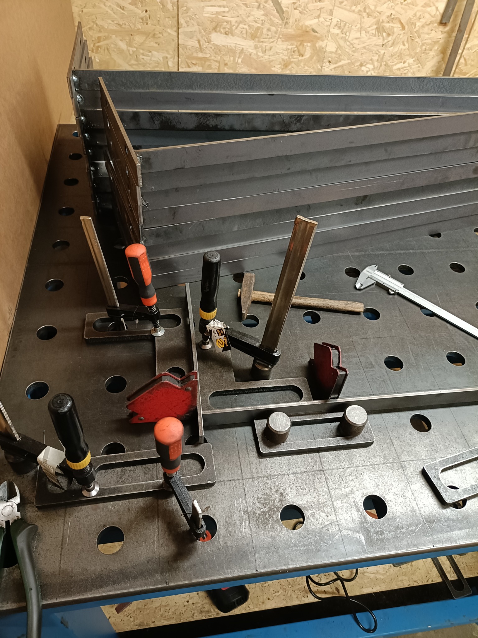 Przygotowane do spawania elementy metalowe, zamocowane na perforowanym stole spawalniczym za pomocą ścisków i kątowników magnetycznych, widoczne narzędzia pomiarowe i młotek.