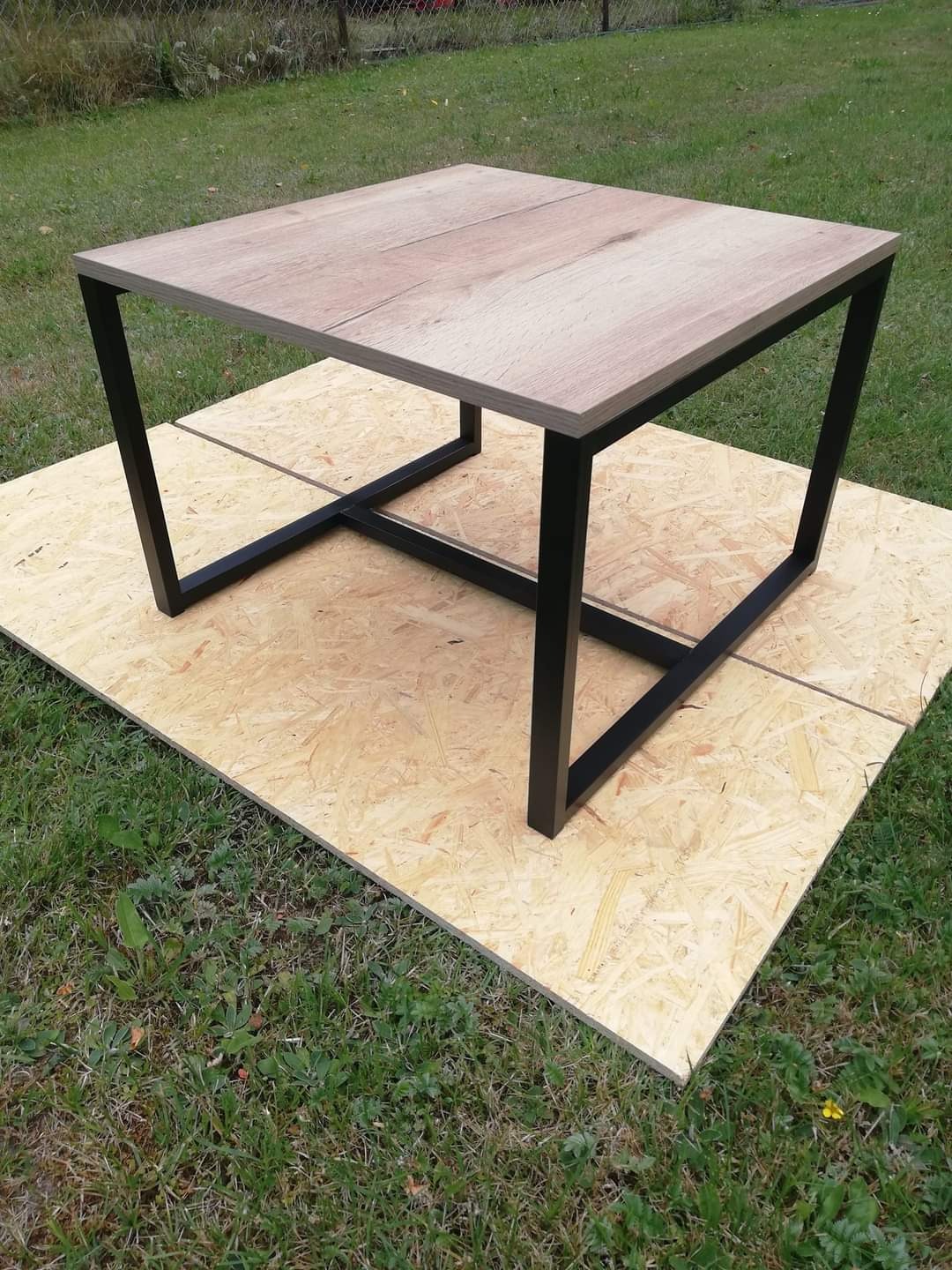 Kwadratowy stolik kawowy z blatem imitującym drewno i czarną metalową podstawą, ustawiony na płycie OSB na trawie.