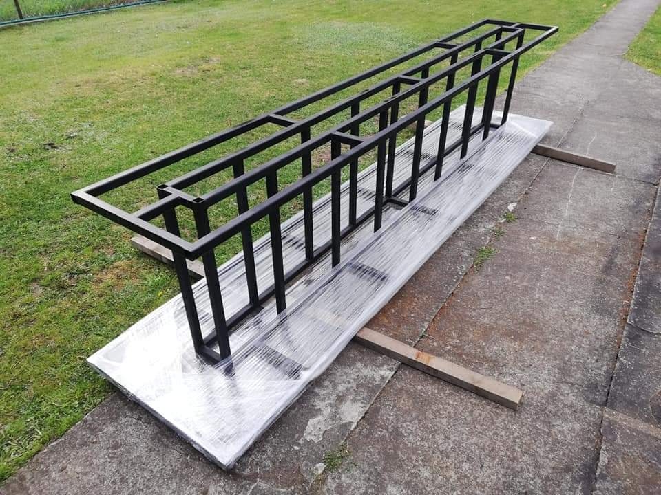 Surowa, czarna, metalowa konstrukcja balustrady, owinięta folią ochronną, leżąca na zewnątrz na betonowej powierzchni z zielonym trawnikiem w tle.