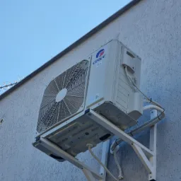 Zewnętrzna jednostka klimatyzacji marki Gree zamontowana na białej ścianie budynku z widocznymi przewodami i wspornikami, na tle błękitnego nieba.