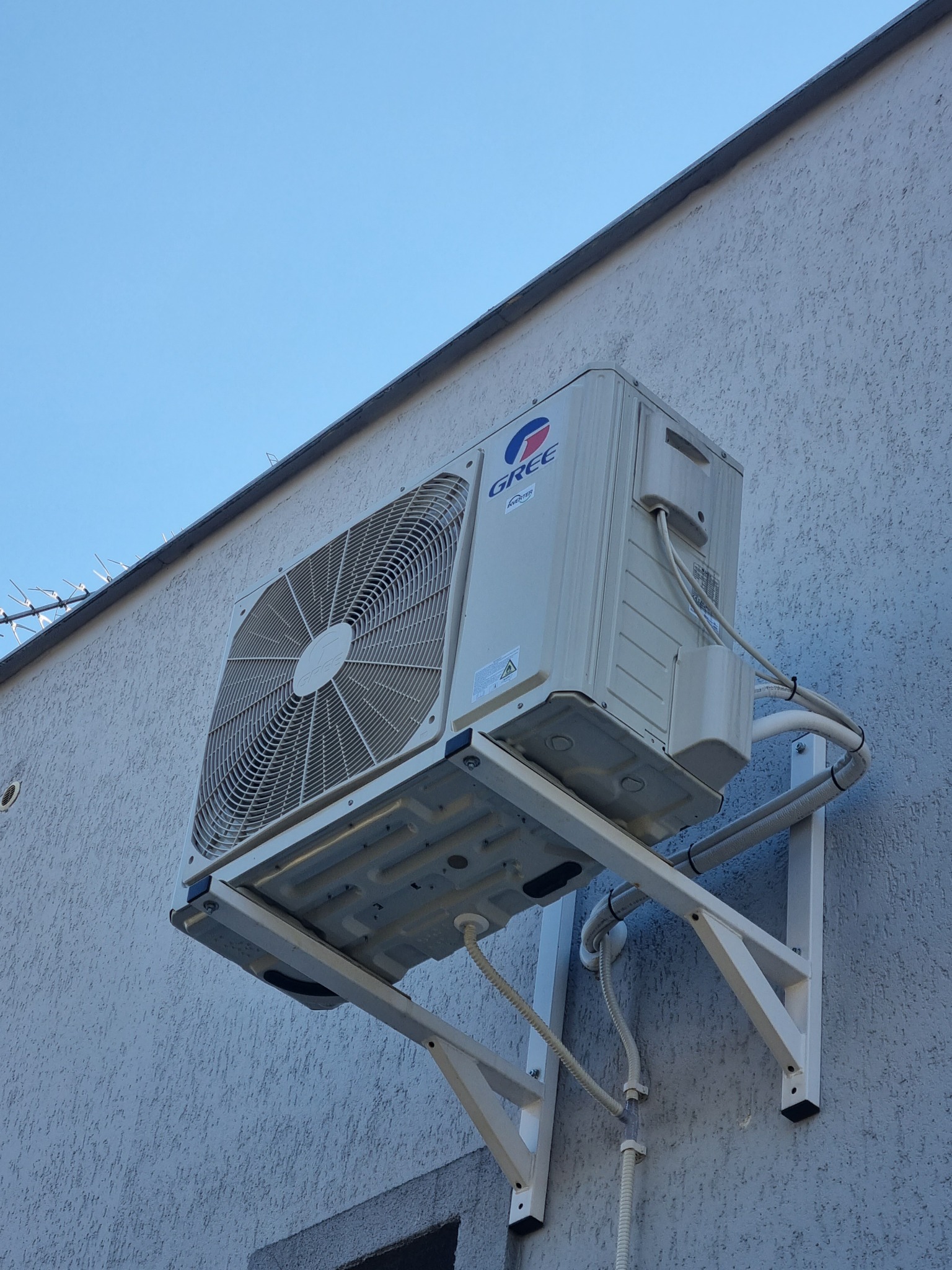 Zewnętrzna jednostka klimatyzacji marki Gree zamontowana na białej ścianie budynku z widocznymi przewodami i wspornikami, na tle błękitnego nieba.