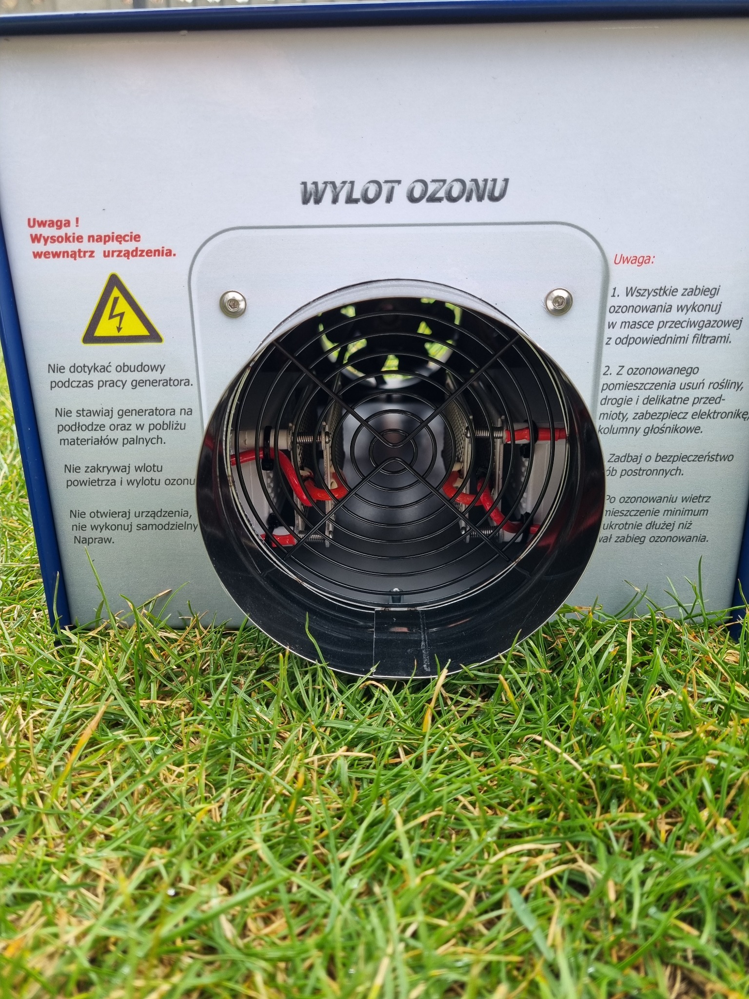 Generator ozonu z widocznym wylotem powietrza i ostrzeżeniami na obudowie, umieszczony na trawie.