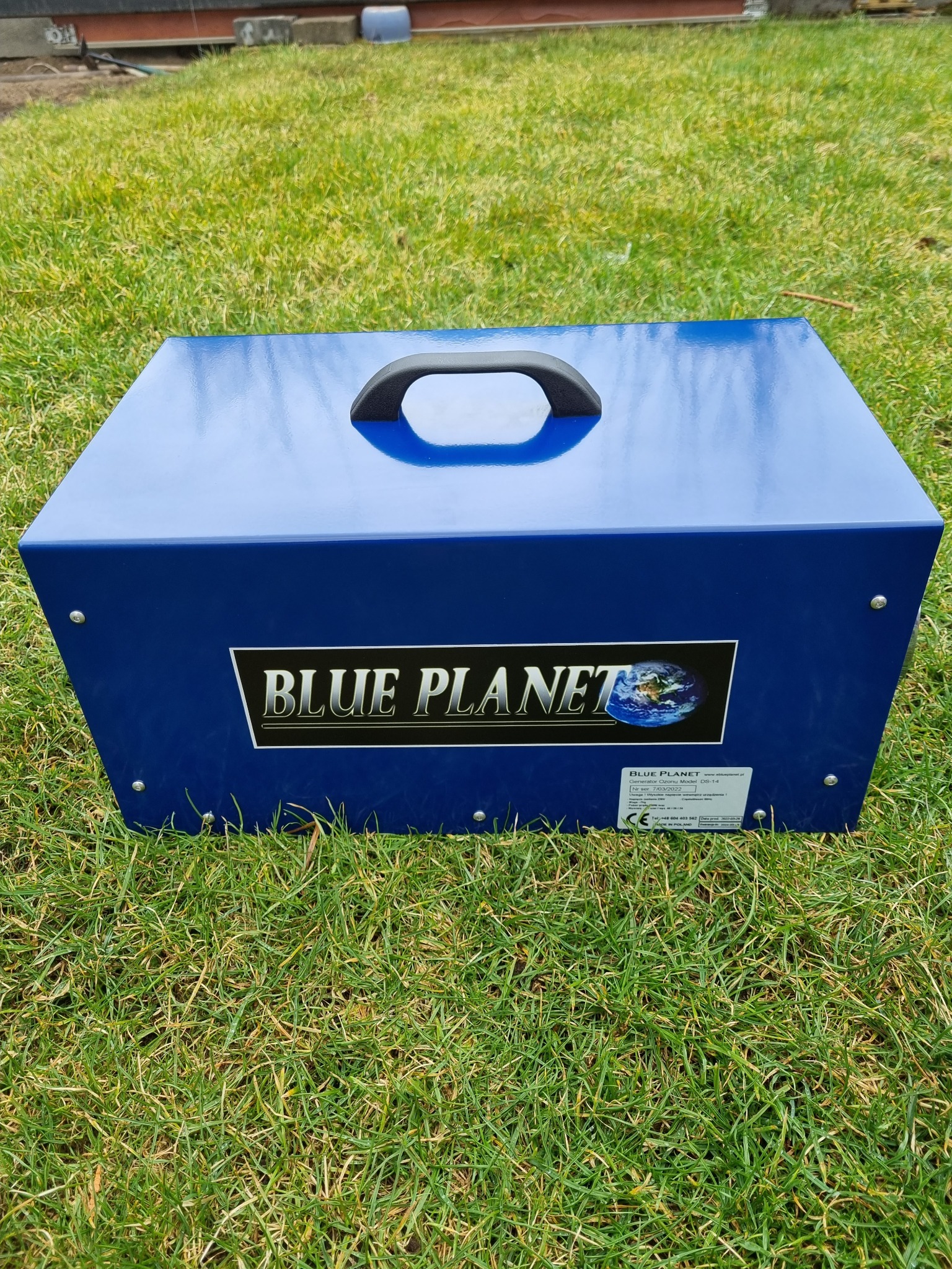 Niebieski generator ozonu Blue Planet DS-14 na trawie, widoczna etykieta z numerem seryjnym i informacjami kontaktowymi.