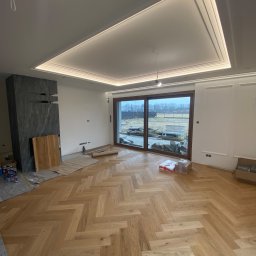 skala - Salon w trakcie wykańczania: parkiet ułożony w jodełkę, widoczne materiały budowlane, nowoczesny kominek z szarego kamienia i drewniane elementy, sufit z oświetleniem LED.