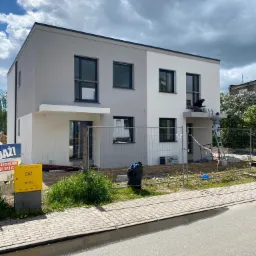 Dwóch mężczyzn maluje elewację dwukondygnacyjnego domu bliźniaczego na biało, stojąc na drabinie i na balkonie, widoczny żółty znak gazowy i ogrodzenie.