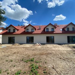 skala - Nowo wybudowany dom jednorodzinny z jasną elewacją i czerwoną dachówką, widok z poziomu gruntu na niezagospodarowanej działce w słoneczny dzień.