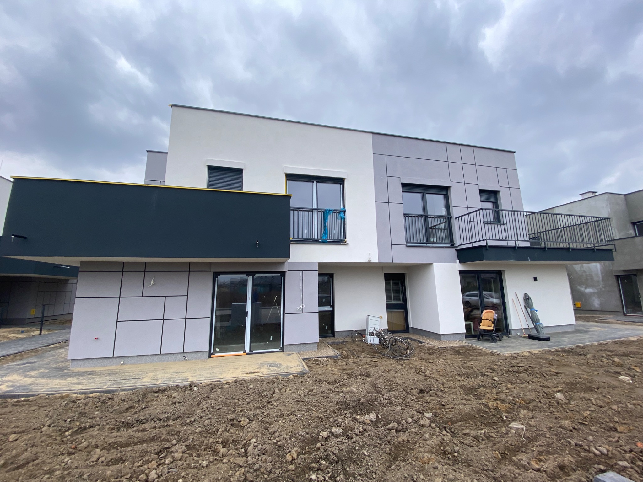 Nowoczesny dom w zabudowie szeregowej we Wrocławiu z elewacją w odcieniach szarości i bieli, częściowo wykończony, z widocznymi śladami prac budowlanych wokół.