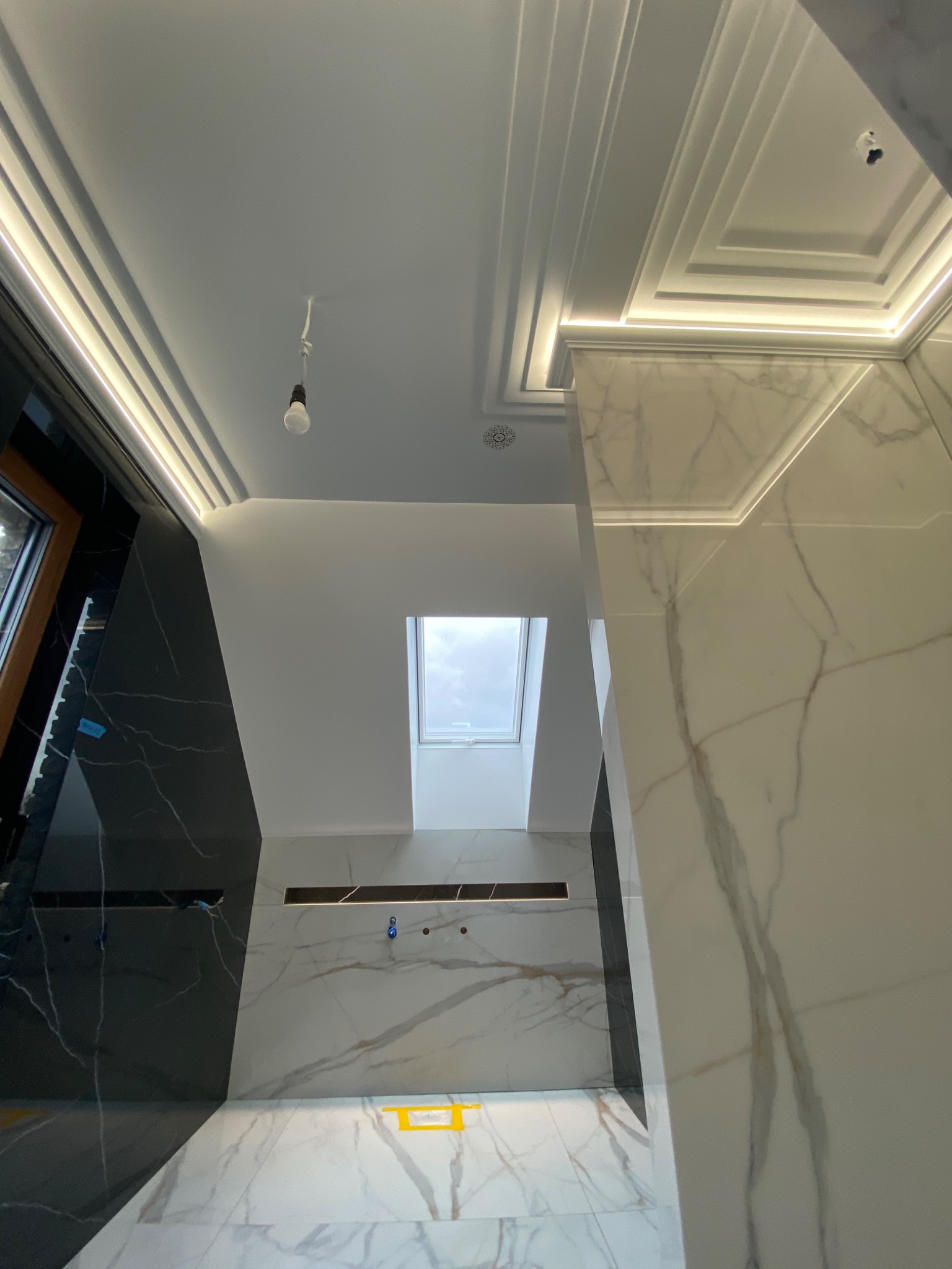 Nowoczesna łazienka w trakcie wykańczania, widoczne marmurowe płytki w odcieniach bieli i czerni, podświetlane sufity kasetonowe i okno dachowe, surowa żarówka zwisająca z sufitu.