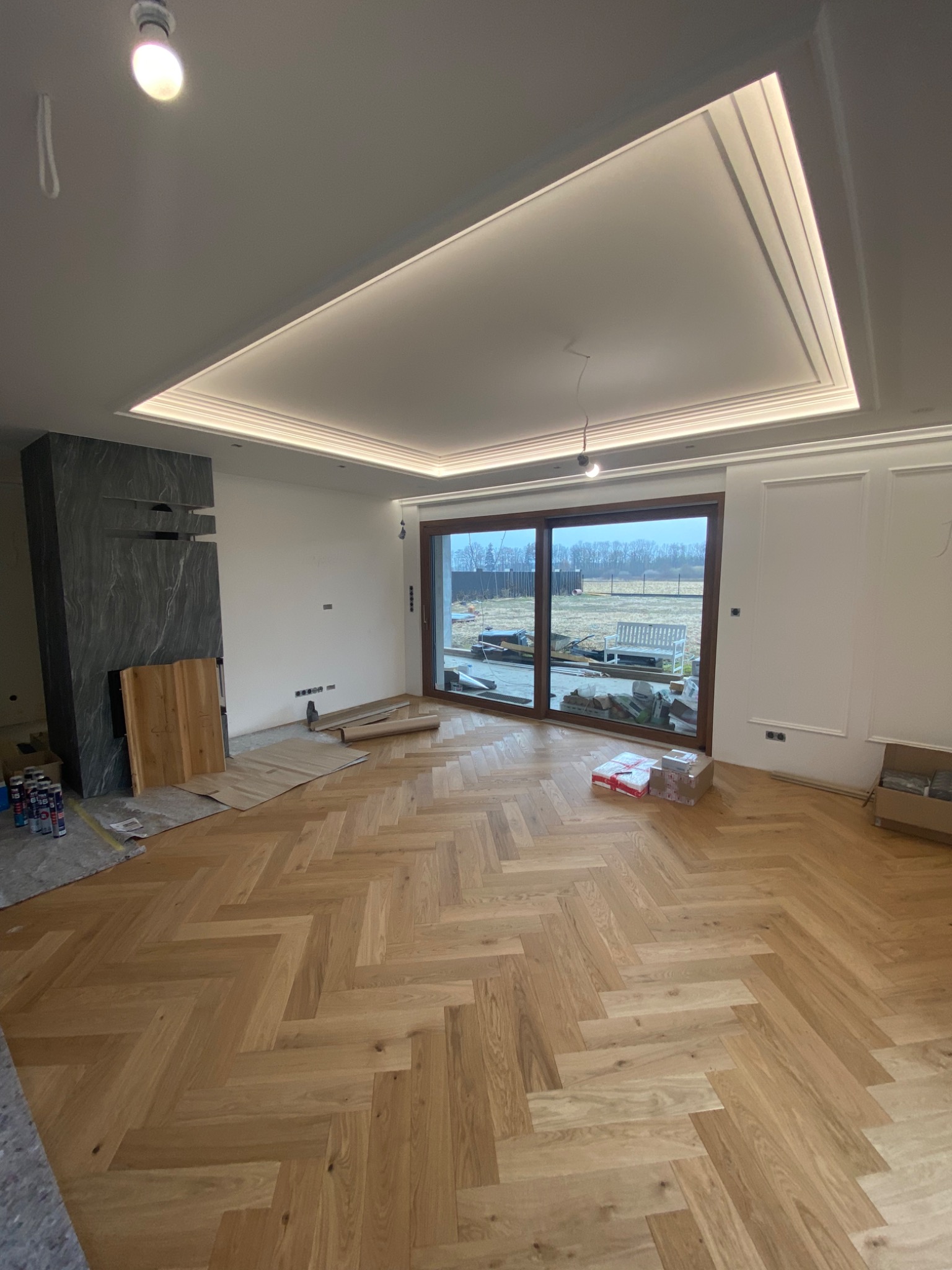 Salon w trakcie wykańczania: parkiet ułożony w jodełkę, widoczne materiały budowlane, nowoczesny kominek z szarego kamienia i drewniane elementy, sufit z oświetleniem LED.