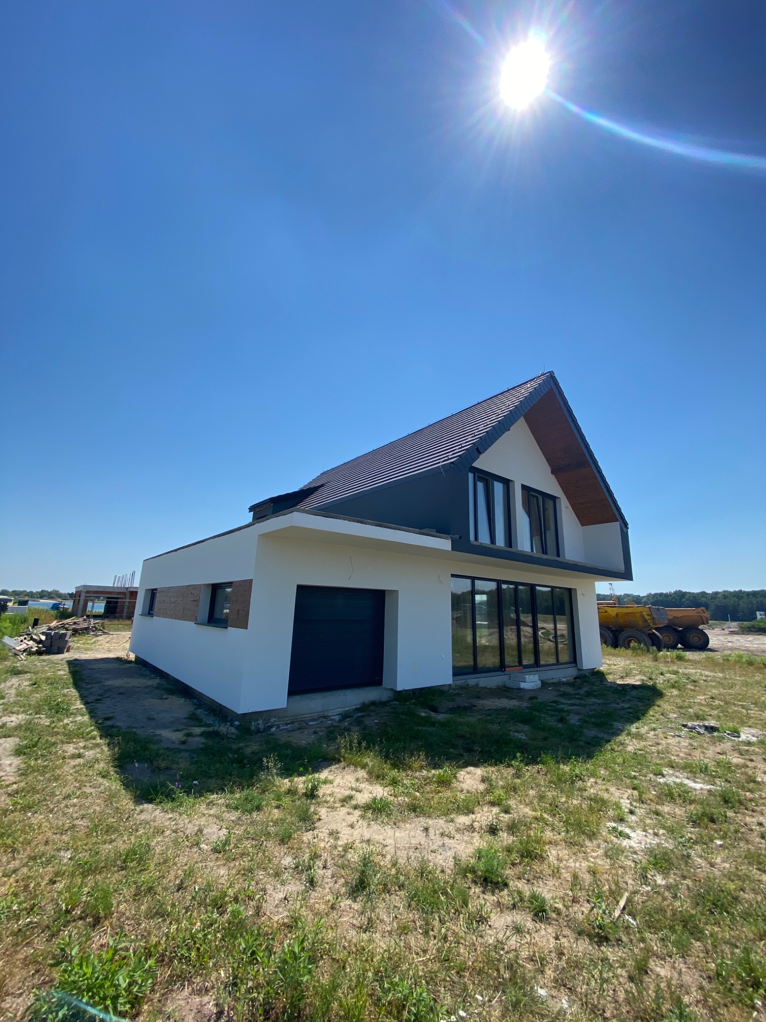 Nowoczesny dom jednorodzinny z płaskim dachem i dużymi przeszkleniami, widoczny na tle błękitnego nieba z intensywnym słońcem, z placem budowy i sprzętem ciężkim w tle.