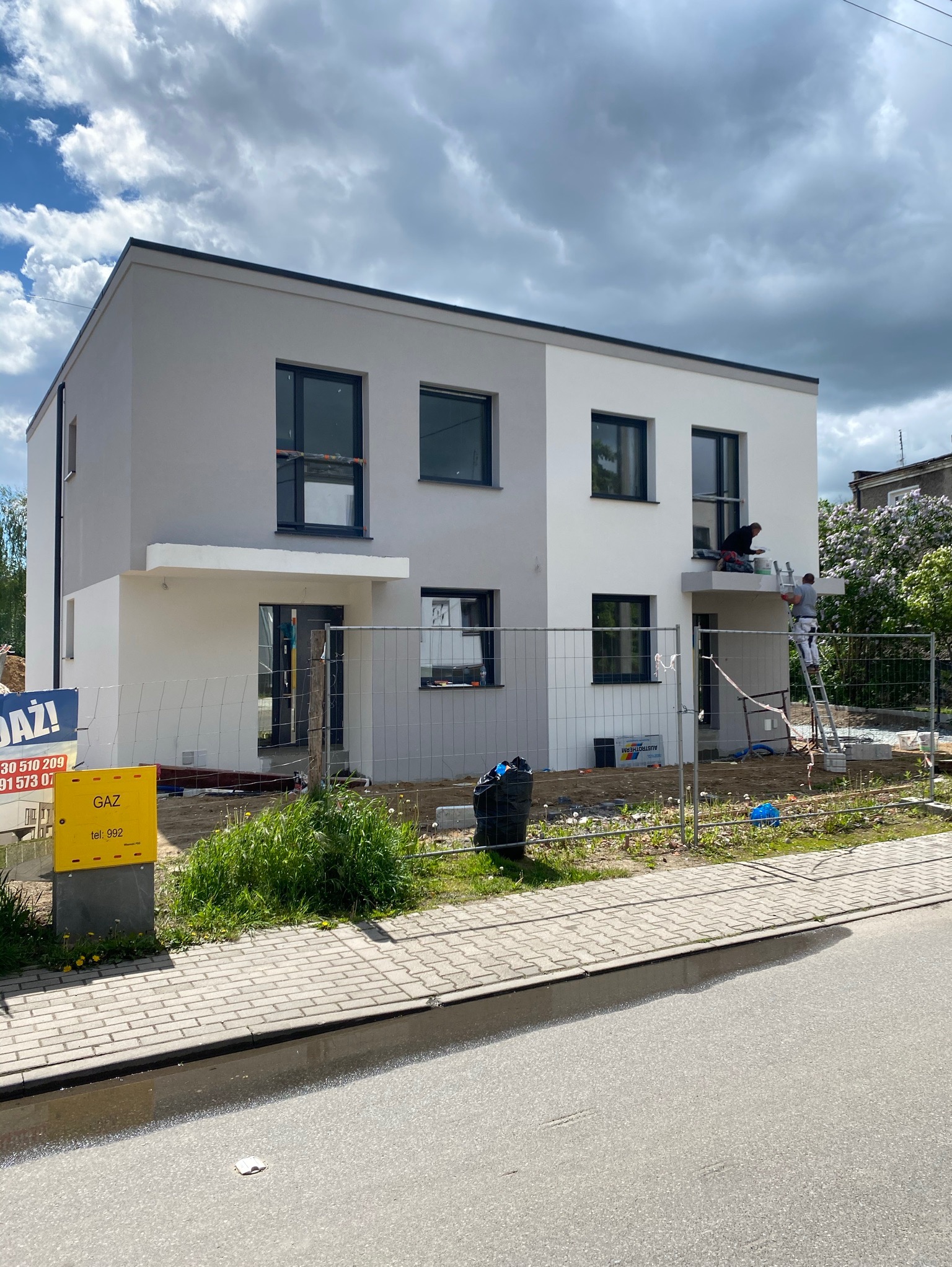 Dwóch mężczyzn maluje elewację dwukondygnacyjnego domu bliźniaczego na biało, stojąc na drabinie i na balkonie, widoczny żółty znak gazowy i ogrodzenie.