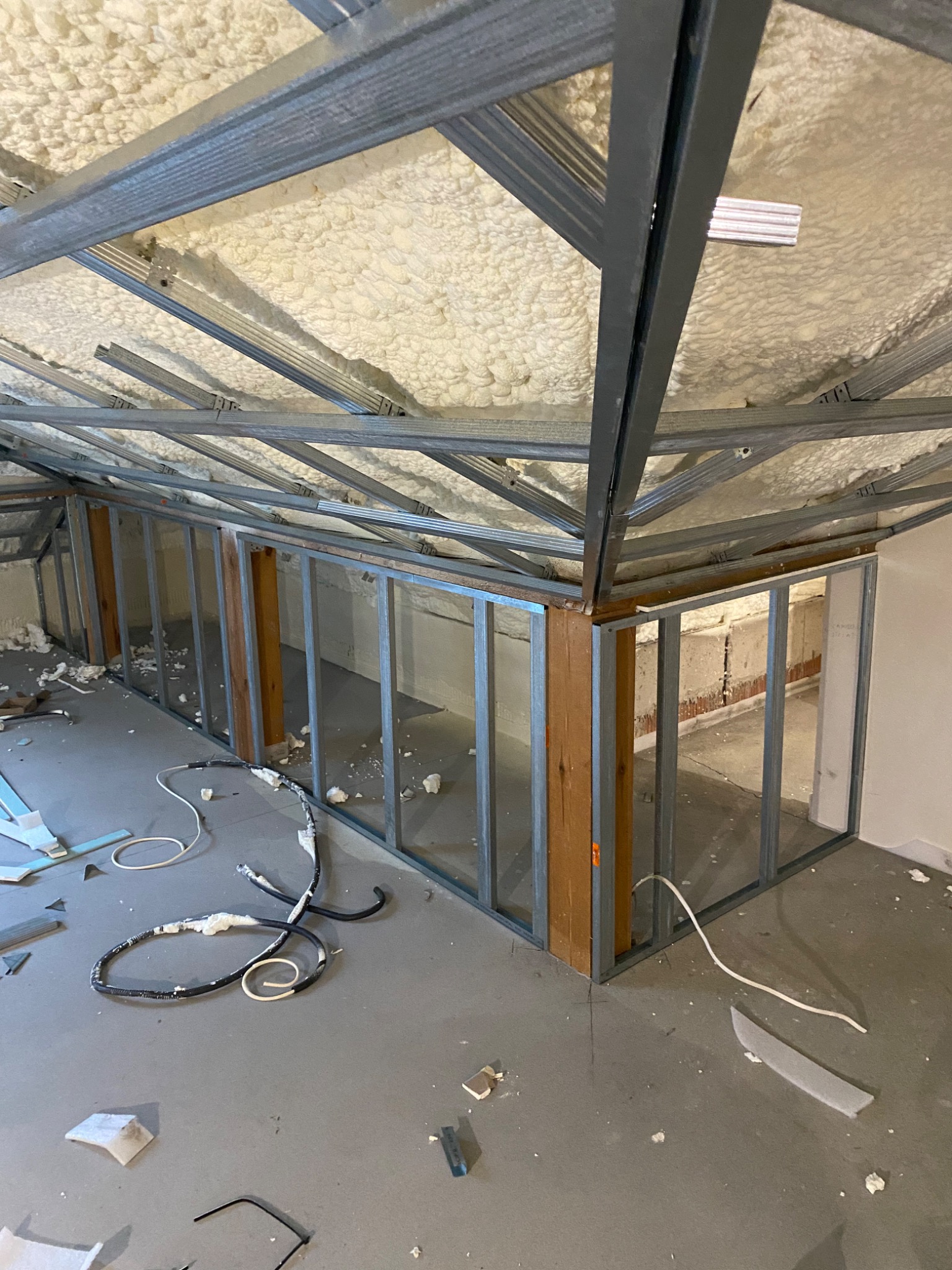 Poddasze w trakcie izolacji pianą, widoczne metalowe profile konstrukcyjne ścianek działowych, drewniane słupy wzmacniające oraz nieuporządkowane przewody i fragmenty materiałów na podłodze.