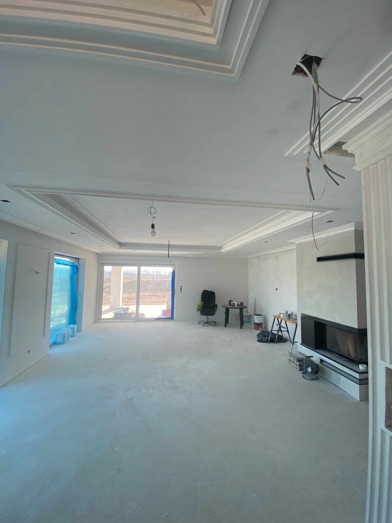 Wnętrze w trakcie wykańczania: surowe ściany przygotowane do malowania, dekoracyjne listwy przysufitowe, częściowo zasłonięte okno folią ochronną, widoczne kable elektryczne zwisające z sufitu...