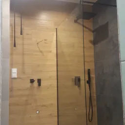 Nowoczesna łazienka z drewnopodobnymi płytkami na ścianie, czarną armaturą i szklaną kabiną prysznicową typu walk-in.