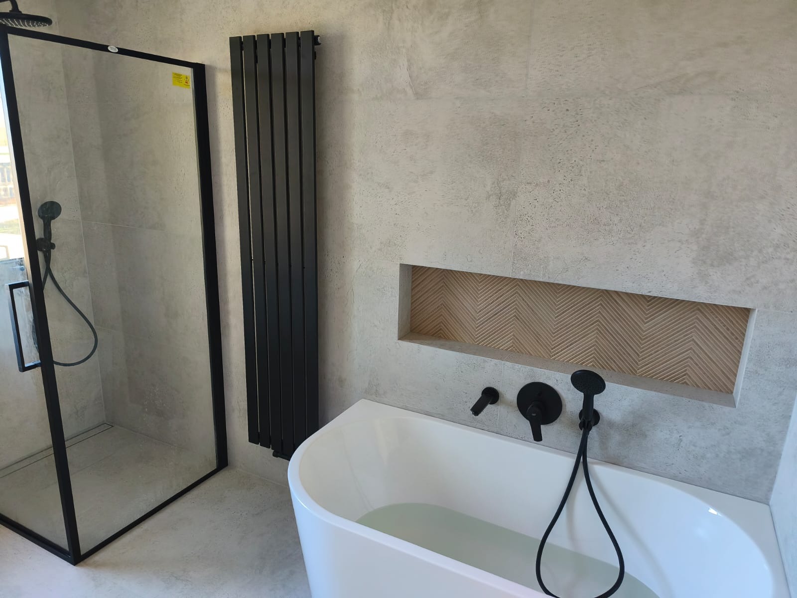 Nowoczesna łazienka z kabiną prysznicową typu walk-in, grzejnikiem drabinkowym i wolnostojącą wanną z czarną armaturą. W ścianie wnęka z dekoracyjnym wykończeniem.