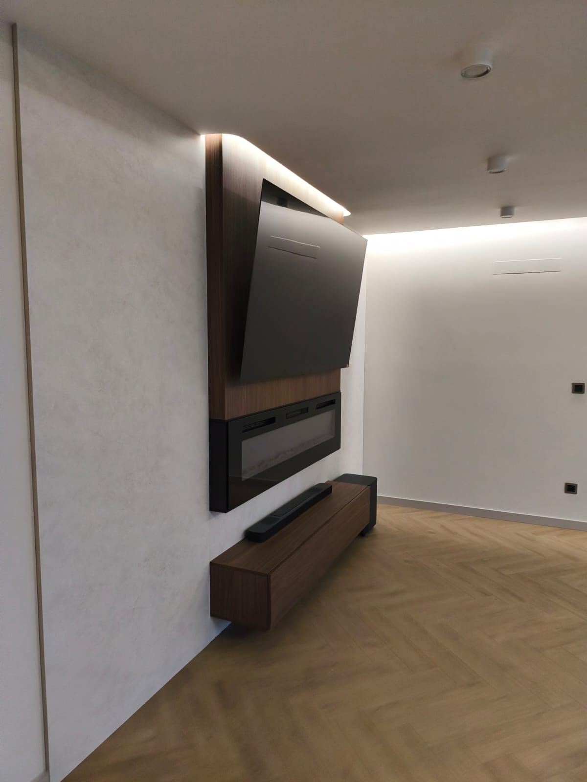 Nowoczesny salon z telewizorem na ścianie z efektem kamienia, podświetleniem LED i drewnianą podłogą w jodełkę. Minimalistyczny design z ukrytymi głośnikami.