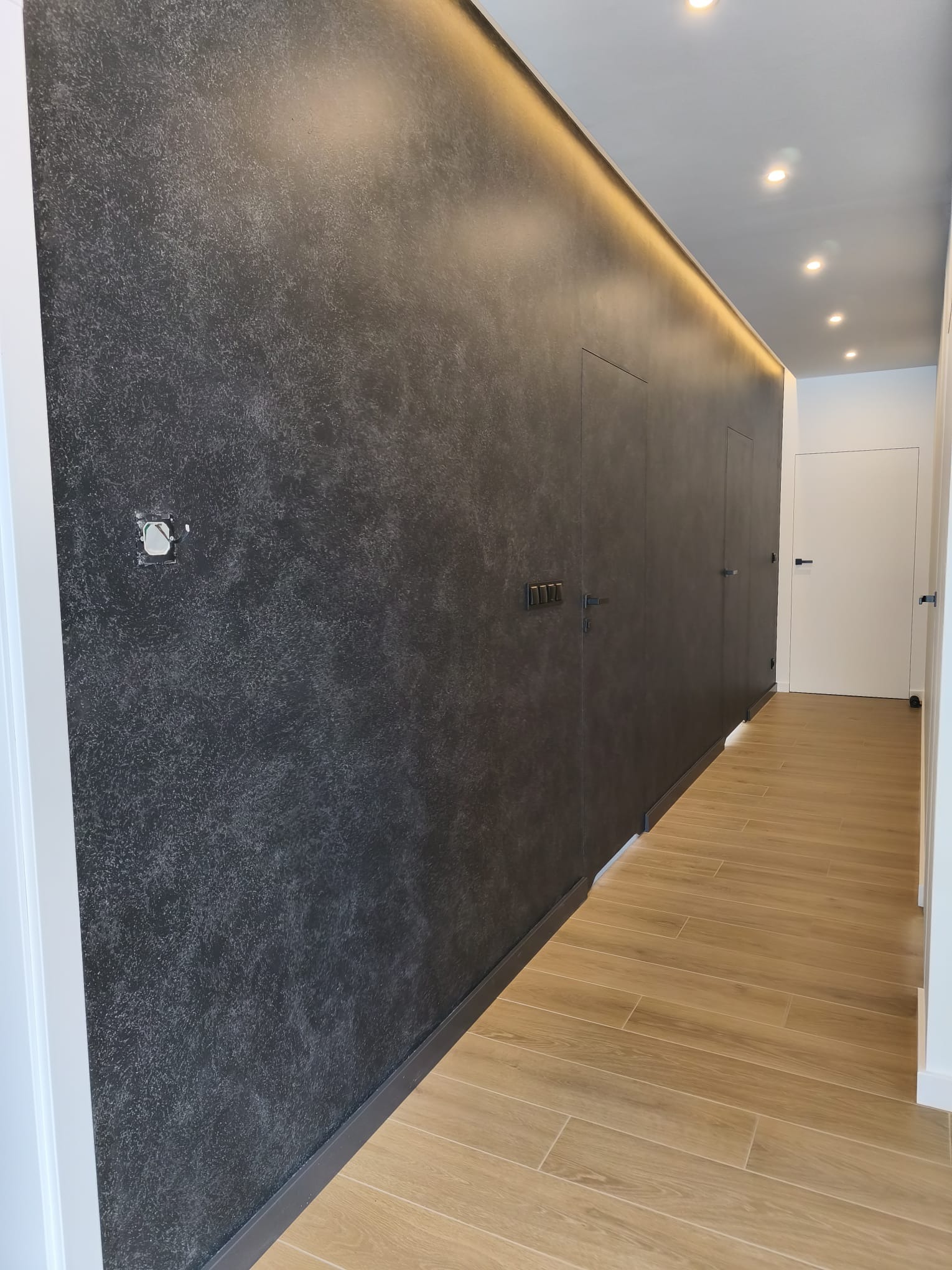 Nowoczesny korytarz po remoncie: ciemna ściana z efektem dekoracyjnym, drewnopodobne płytki na podłodze, ukryte drzwi i oświetlenie LED wzdłuż sufitu.