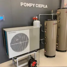 Wewnętrzna instalacja z pompą ciepła Technix, dwa pionowe zbiorniki w kolorze taupe, ekspozycja w szarym pomieszczeniu z napisem 'Pompy ciepła' na ścianie.