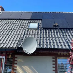 Czyszczenie paneli słonecznych na ciemnym dachu z dachówki przy pomocy szczotki na teleskopowym kiju, widoczny fragment domu z anteną satelitarną i czerwonym drzewem.