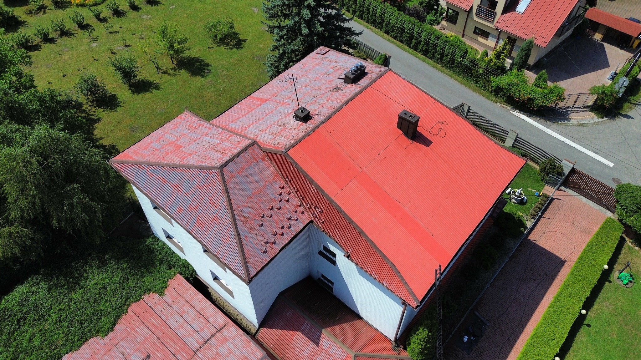 Czerwony dach domu z blachy trapezowej, widok z lotu ptaka. Część dachu pomalowana, część z widocznymi śladami korozji. Kominy i anteny na dachu. Otoczenie: zieleń, zabudowa jednorodzinna.