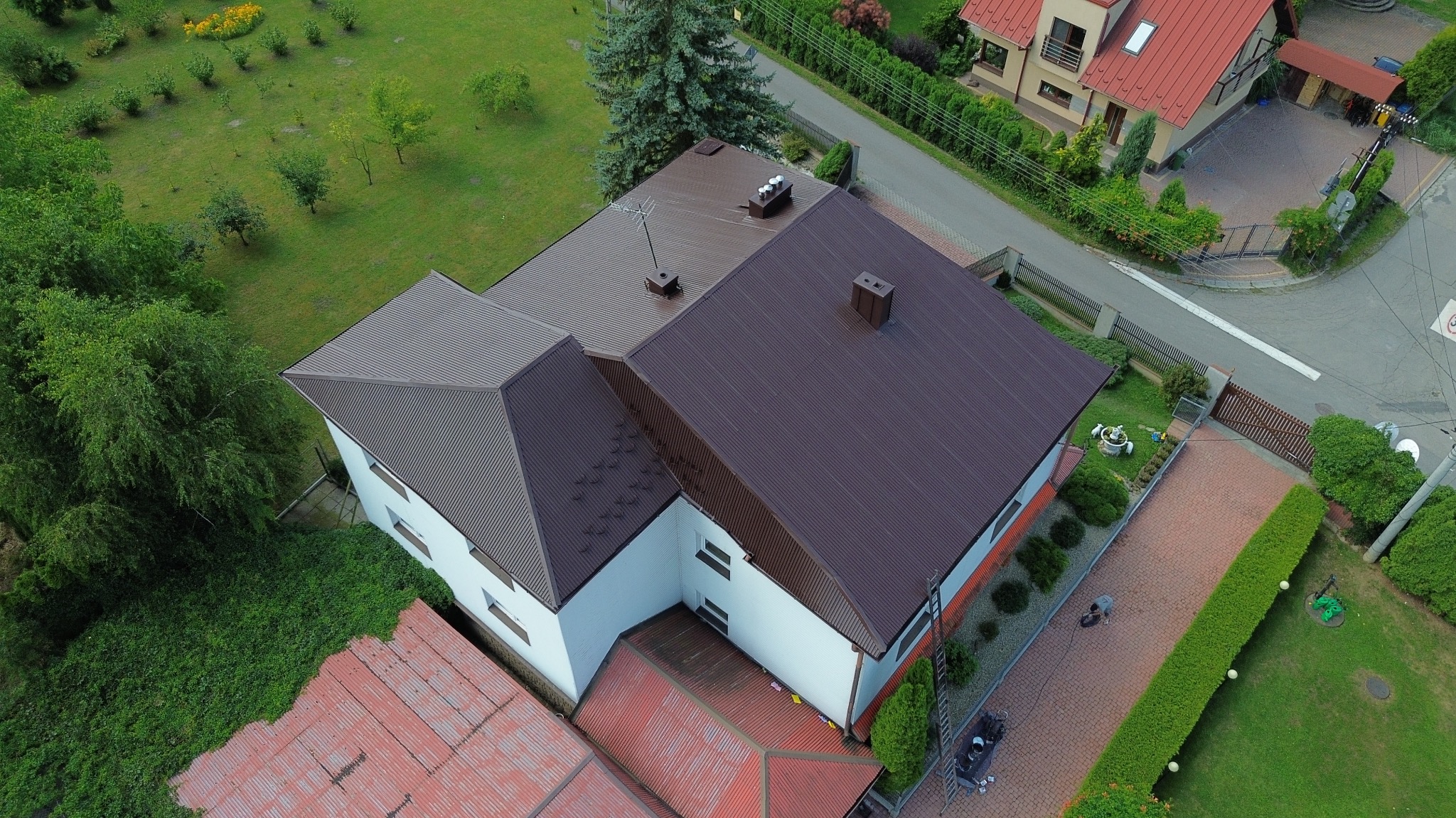 Zdjęcie z drona przedstawia dom z brązowym dachem z blachy trapezowej, białą elewacją i zielonym otoczeniem. Widoczny komin, drzewa i fragment ulicy.