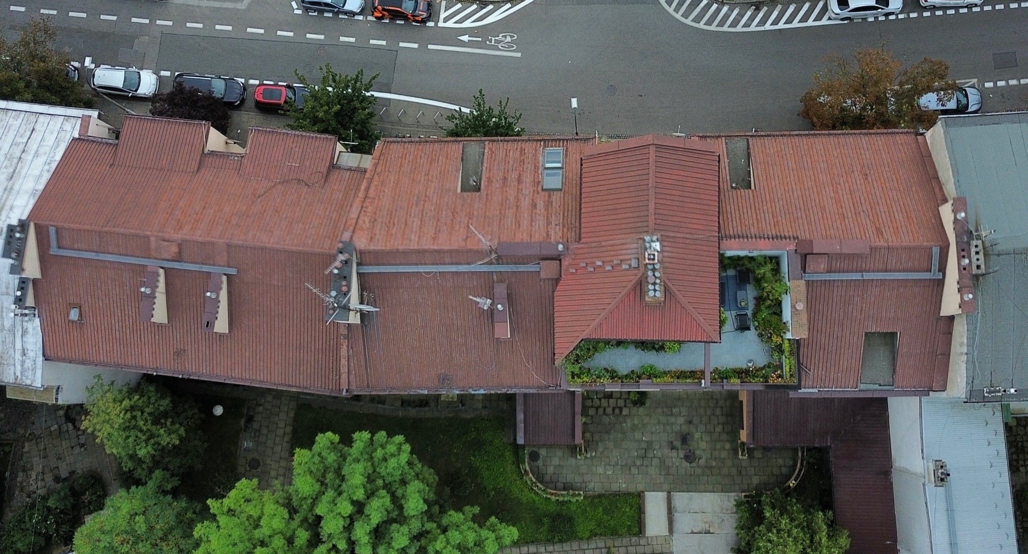 Widok z góry na budynek z czerwonym dachem, z widocznymi elementami architektury, zielenią na dachu i w ogrodzie, oraz fragmentem ulicy z zaparkowanymi samochodami.