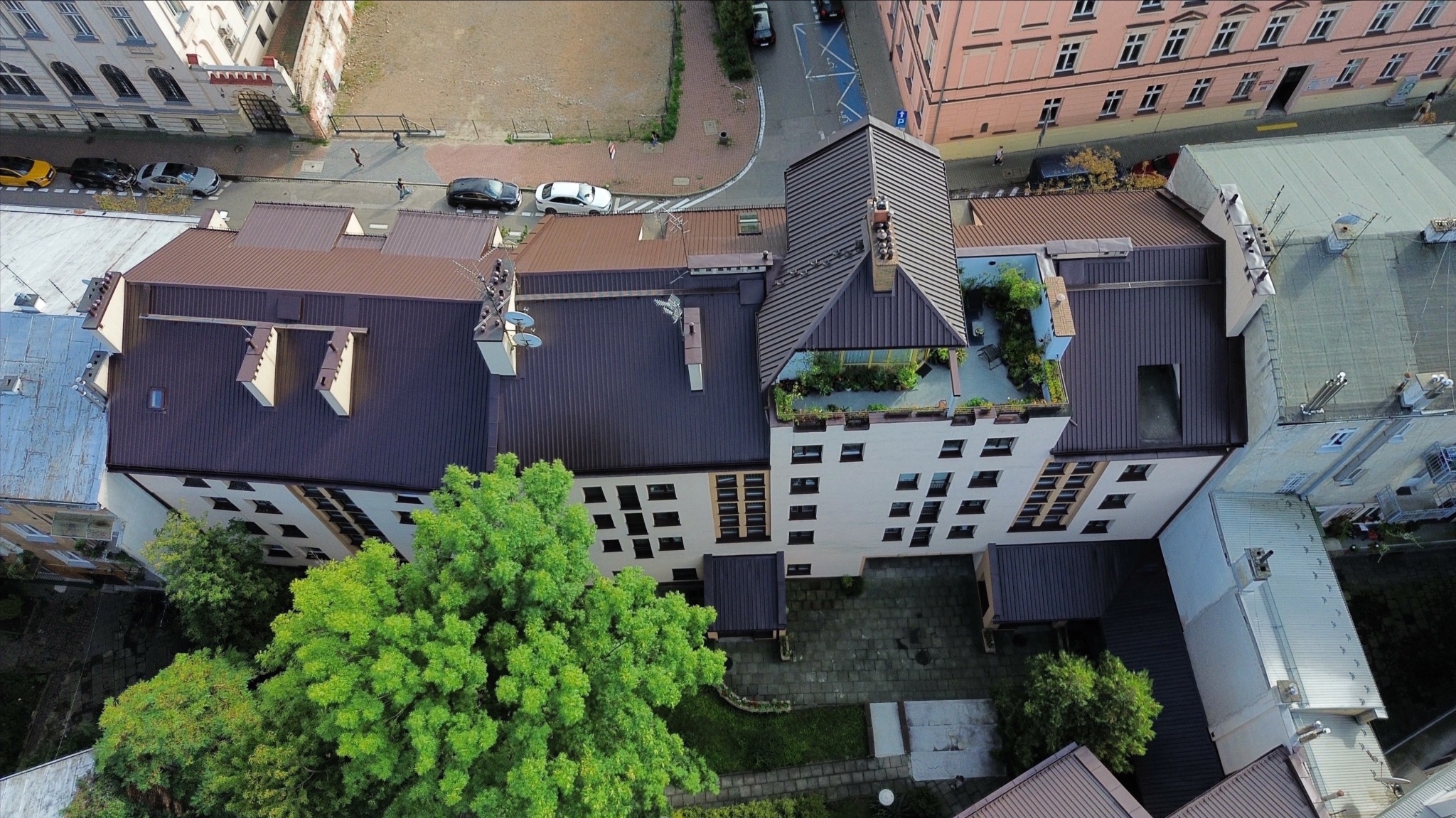 Widok z góry na budynek z ciemnym dachem i jasną elewacją, otoczony zielenią i innymi budynkami w miejskiej scenerii. Dach z elementami dekoracyjnymi i roślinnością.