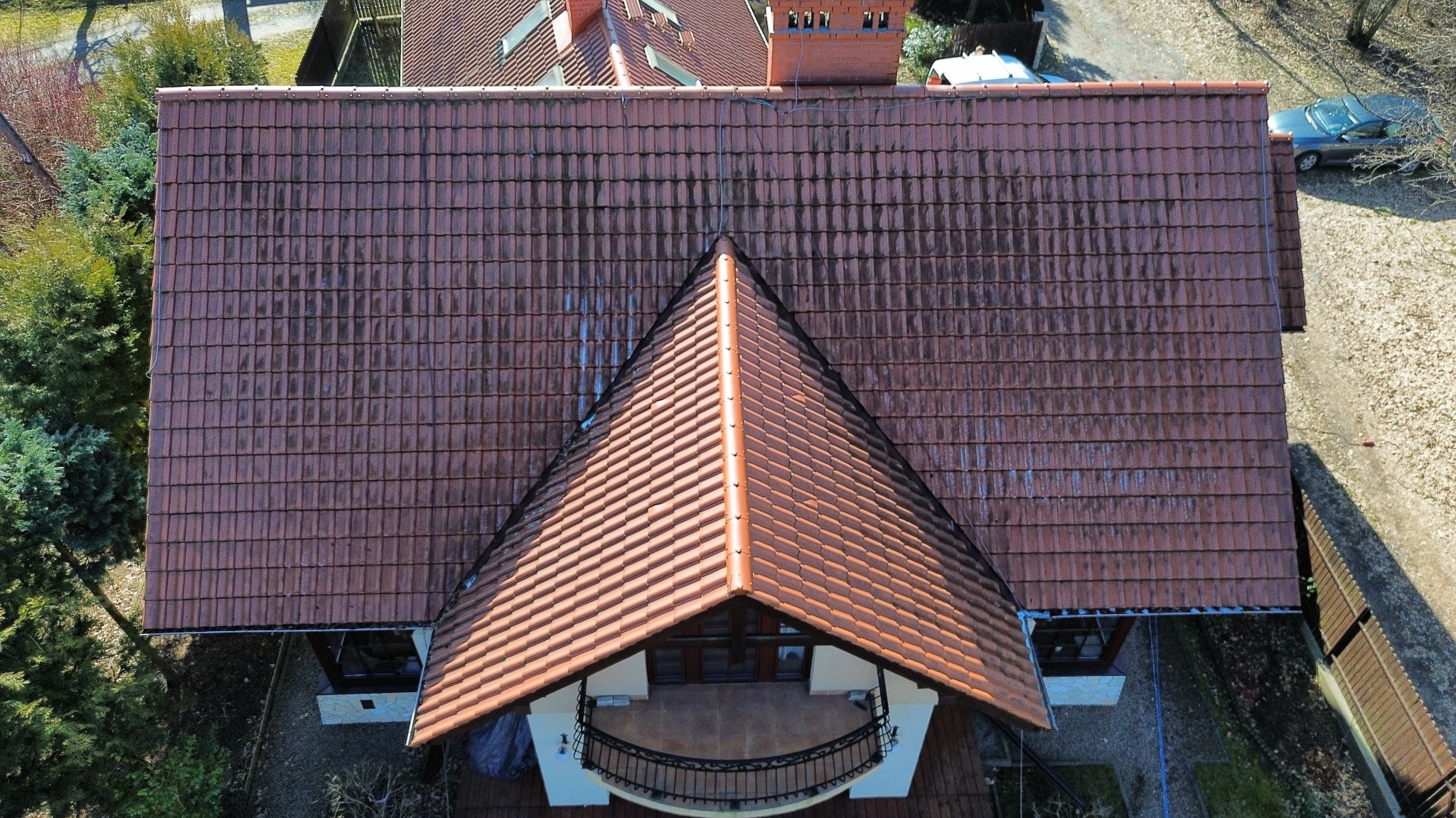 Czerwona dachówka ceramiczna na dachu domu z widocznymi śladami zabrudzeń, sfotografowana z góry, z widocznym fragmentem balkonu i otoczenia.