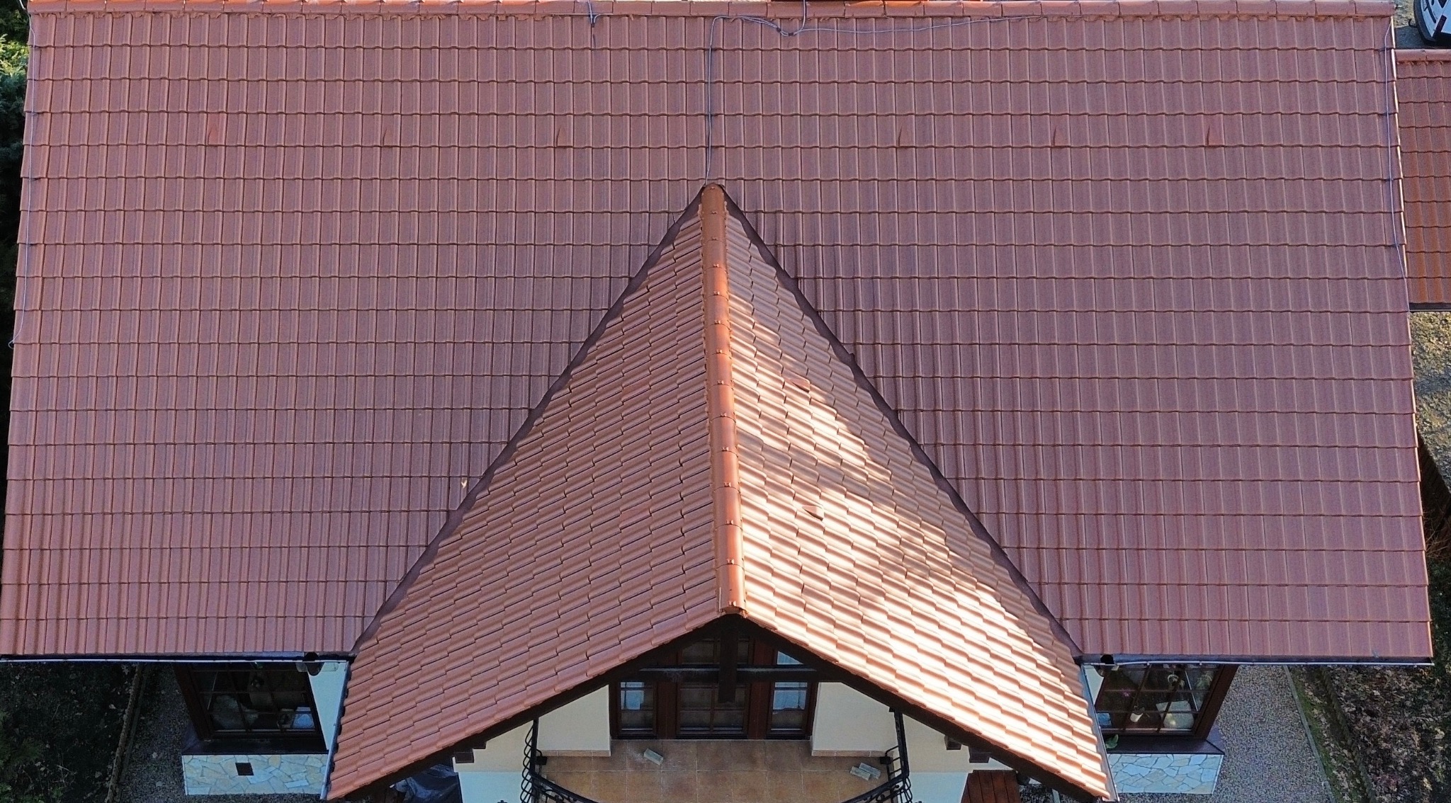Czerwona dachówka ceramiczna na dachu domu, widok z góry. Regularny wzór ułożenia dachówek, symetryczny kształt dachu. Częściowo widoczny fragment elewacji budynku.