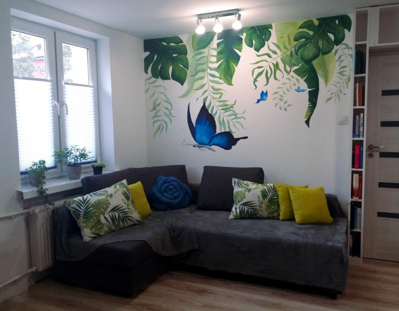 Pokój z narożną sofą, poduszkami w tropikalne wzory i żółtymi, dekoracyjną poduszką w kształcie niebieskiej róży, ścianą z ręcznie malowanymi motywami roślinnymi i motylami, oraz regałem na książki