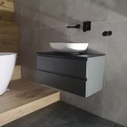 Nowoczesna łazienka z wiszącą szafką pod umywalkę w kolorze grafitowym, białą umywalką nablatową i czarną armaturą na tle szarej ściany z marmurkowym wzorem, obok widoczna biała wanna wolnostojąca.