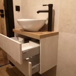 Nowoczesna łazienka: biała umywalka nablatowa z czarną armaturą na szafce z szufladami Blum, widoczna faktura okładziny meblowej i płytek ściennych.