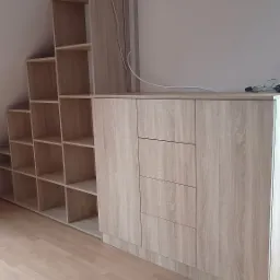 Zabudowa pod skosem z jasnego drewna: regał schodkowy z otwartymi półkami połączony z komodą z trzema szufladami i szafkami, widoczne okablowanie na górze.